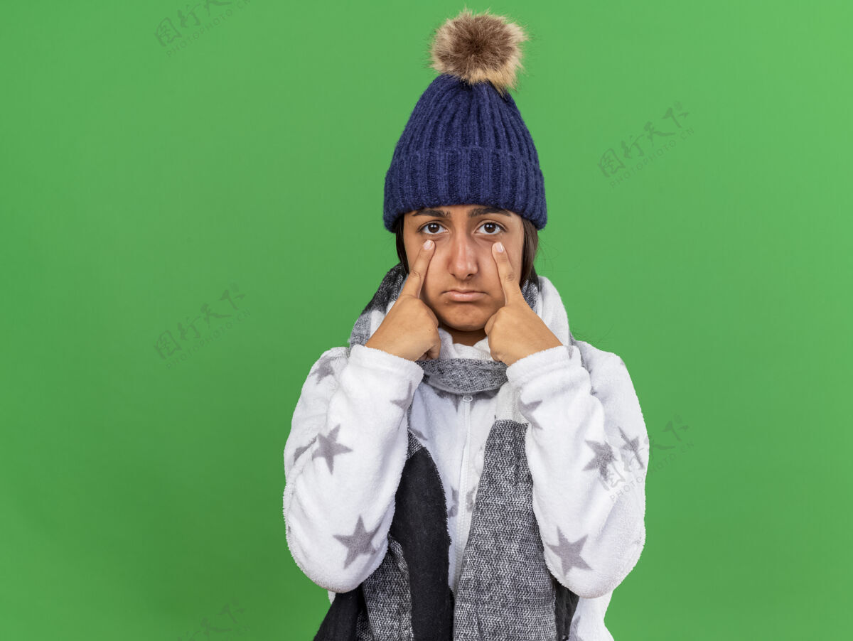 帽子悲伤的生病的年轻女孩戴着冬天的帽子 戴着围巾 手指放在绿色背景上孤立的眼睛上悲伤眼睛疾病