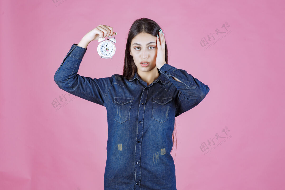 女人穿着牛仔衬衫的年轻女子拿着闹钟 因为戒指而捂着耳朵噪音年轻人休闲
