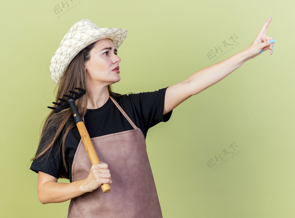 女孩看着一边迷茫美丽的园丁女孩穿着制服戴着园艺帽把耙子放在肩上 指着一边孤立的橄榄绿背景制服点混乱
