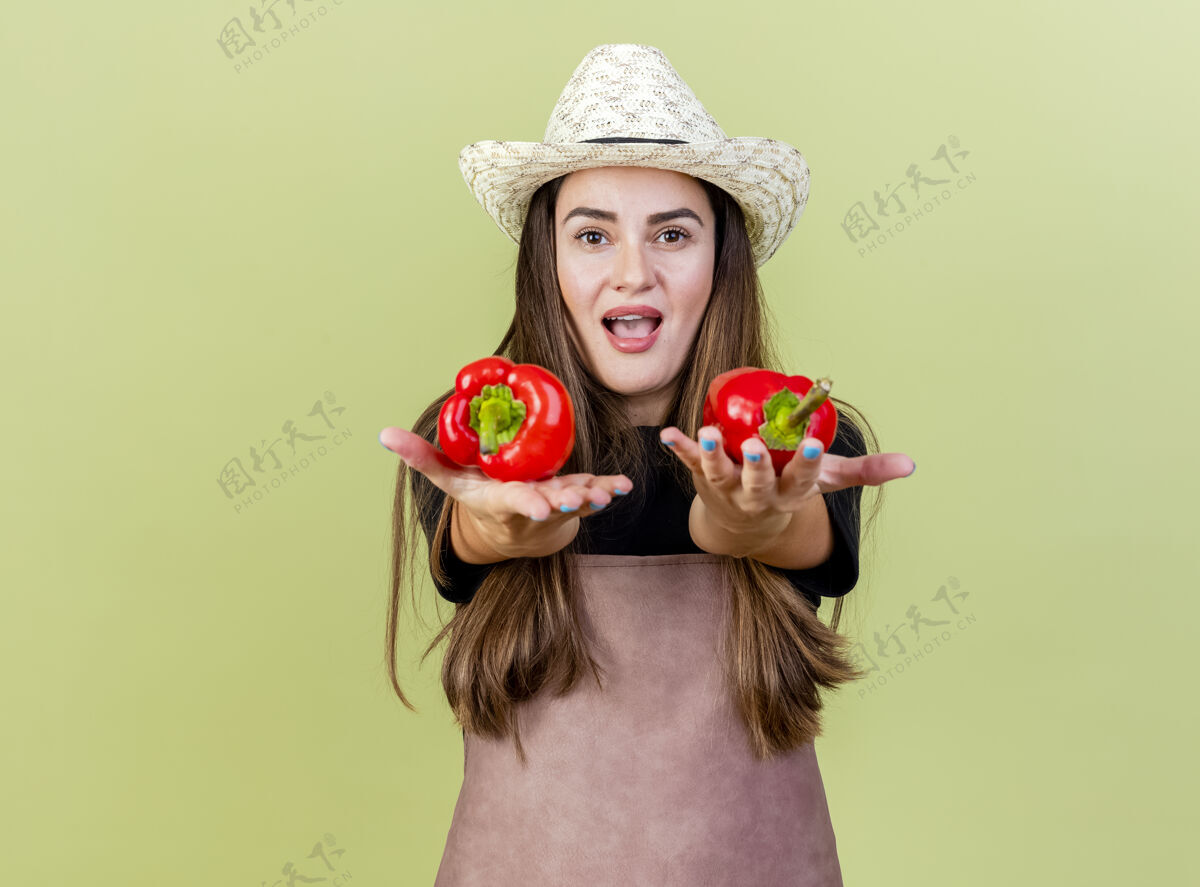 橄榄身着制服的快乐美丽的园丁女孩戴着园艺帽 在橄榄绿的背景下 对着镜头伸出辣椒花园胡椒相机