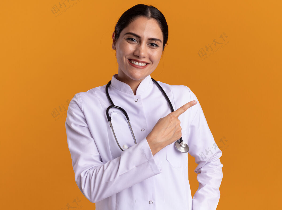 外套年轻的女医生穿着医用外套 脖子上戴着听诊器 微笑着自信地看着前面 用食指指着站在橙色墙上的一边信心听诊器医生