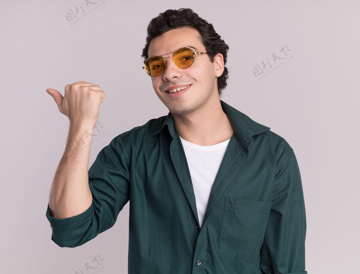 衬衫穿绿衬衫的年轻人戴着眼镜 面带微笑 用拇指指着前面站在白墙上微笑戴着脸