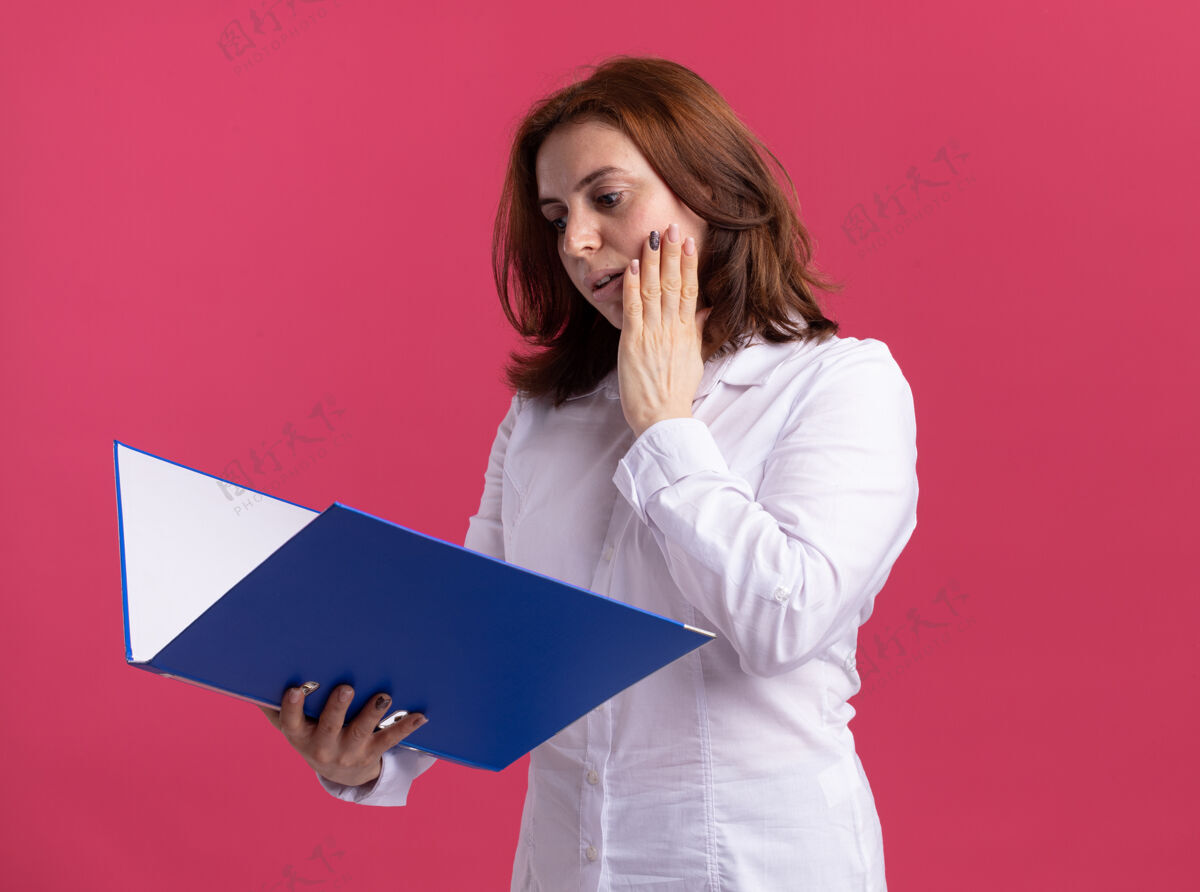 年轻穿着白衬衫的年轻女子拿着文件夹 困惑地看着它站在粉红色的墙上女人站夹