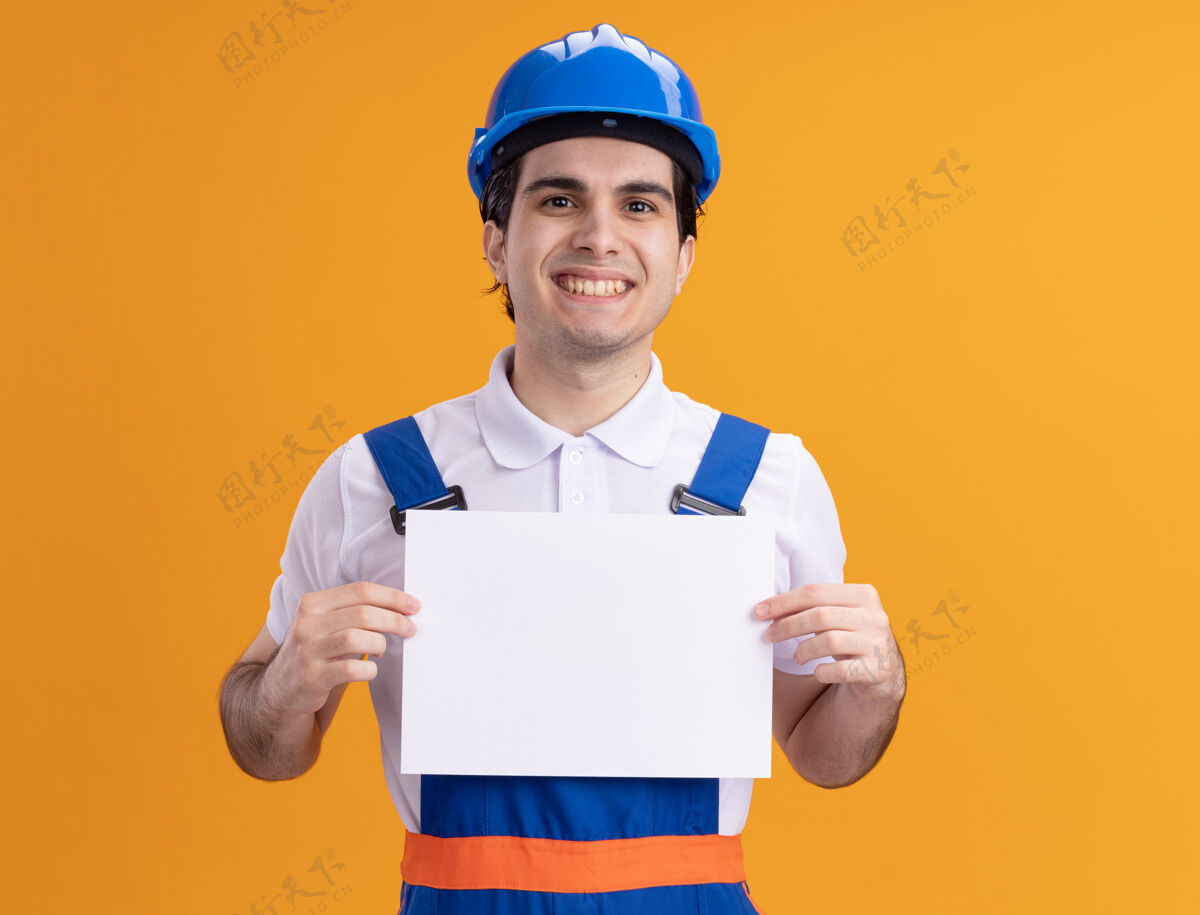 空白年轻的建筑工人身穿建筑制服 戴着安全帽 手里拿着空白的一页 站在橙色的墙上 面带微笑地看着前面制服微笑举行