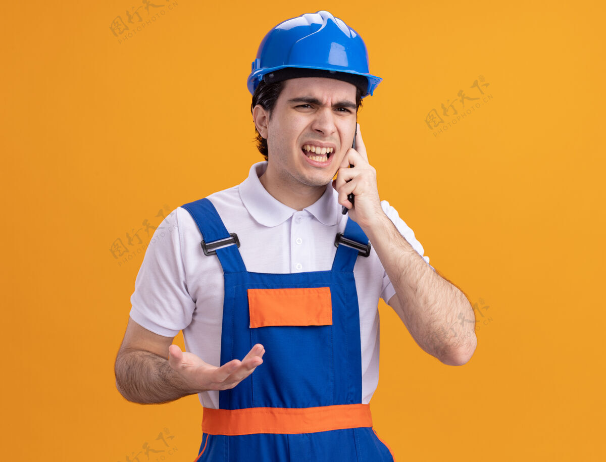 制服愤怒的年轻建筑工人穿着建筑制服 戴着安全帽 站在橙色的墙上讲着手机头盔建筑工人年轻