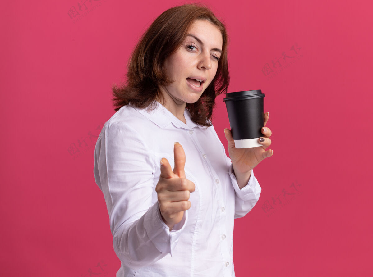 快乐穿着白衬衫的快乐的年轻女子拿着咖啡杯 食指指着前面 微笑着 眨着眼睛站在粉红色的墙上咖啡杯子眨眼