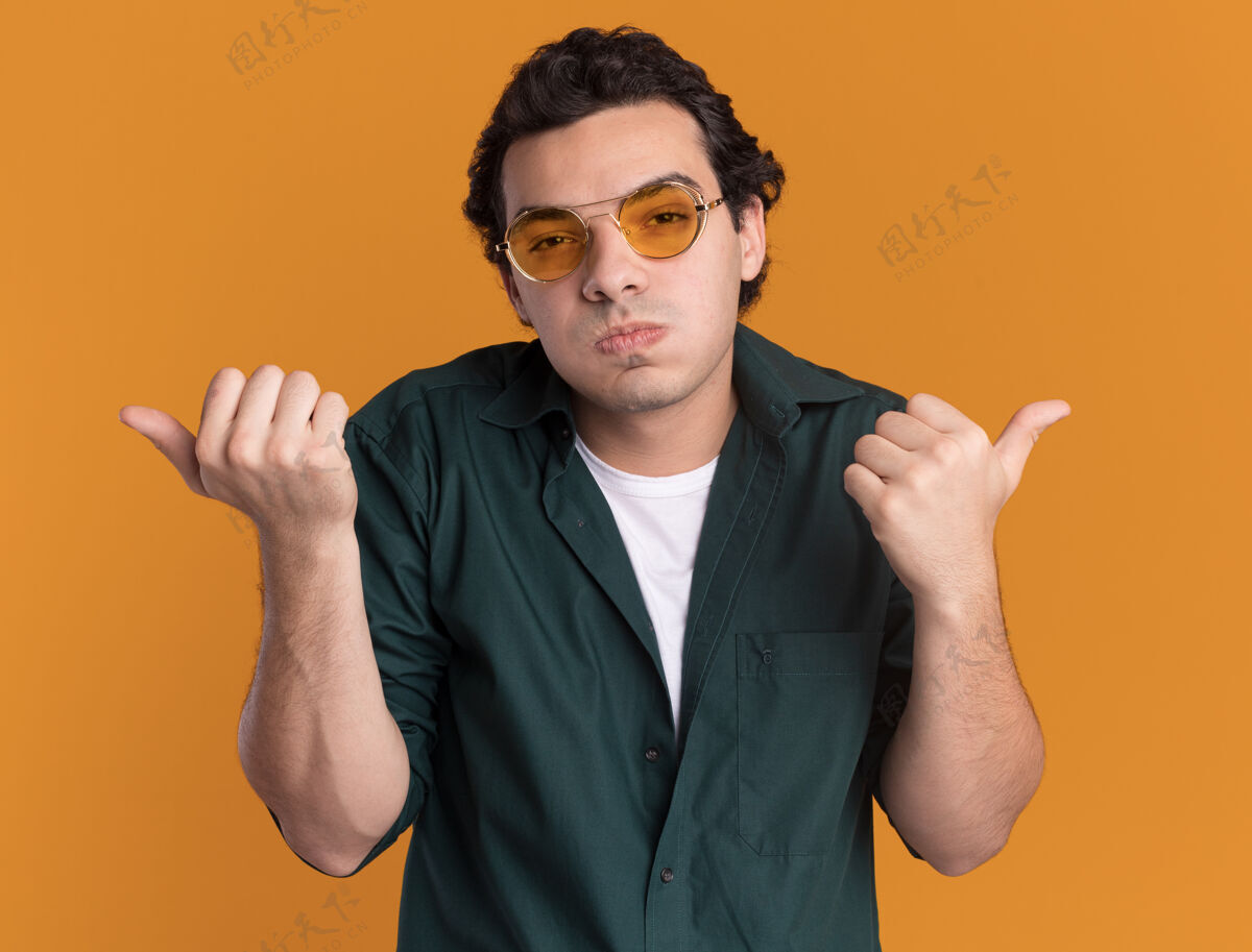 脸颊身穿绿衬衫的年轻人戴着眼镜 困惑地看着前面吹着脸颊 用大拇指指着两边站在橘色的墙上穿指点衬衫