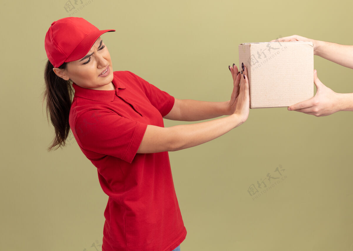 帽子身穿红色制服 戴着帽子的年轻送货员站在绿色的墙上拒绝拿箱子盒子制服送货