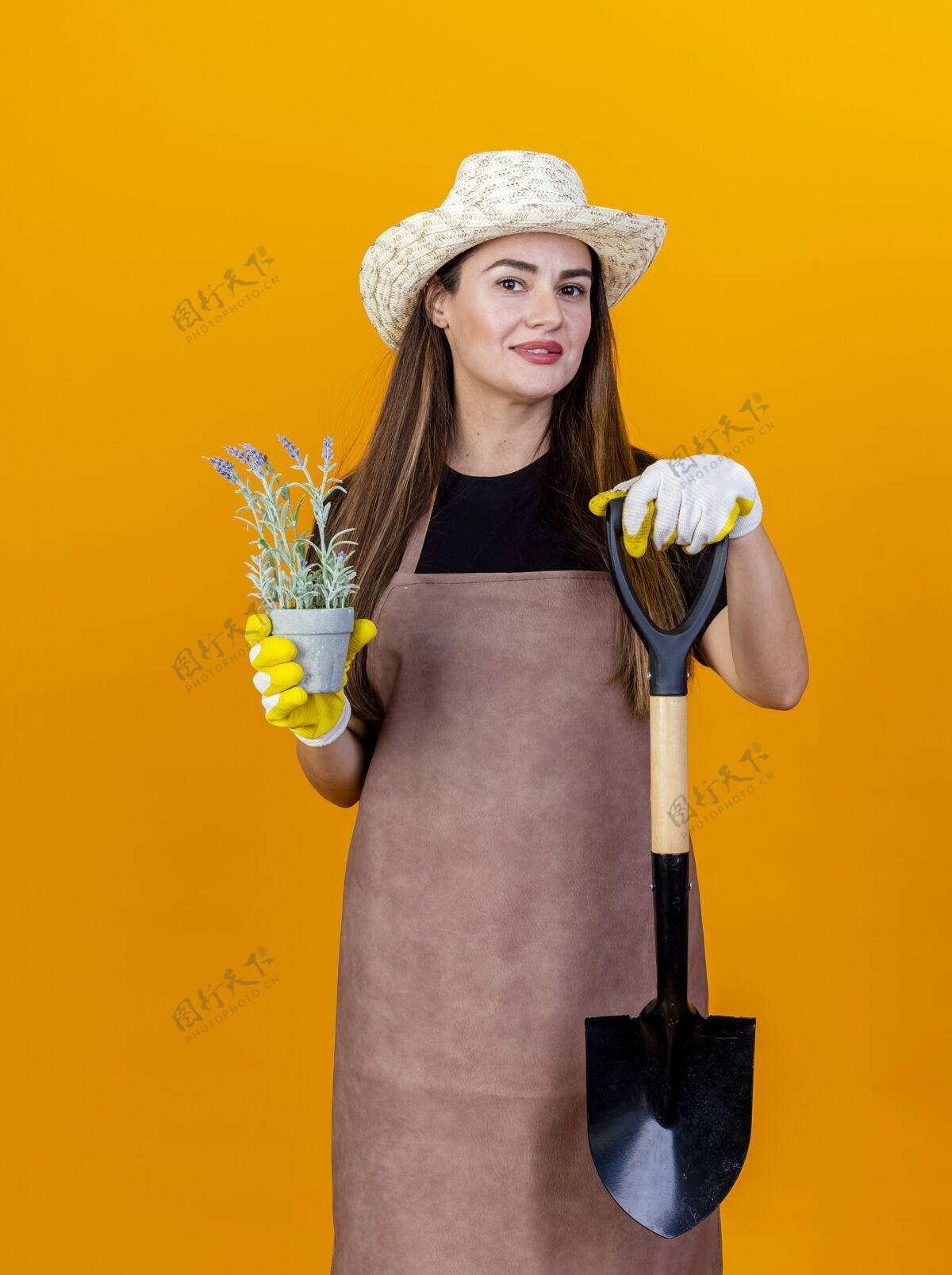 帽子微笑美丽的园丁女孩穿着制服 戴着园艺帽 戴着手套 拿着铲子 花盆里的花被隔离在橙色的背景上花盆手拿园艺