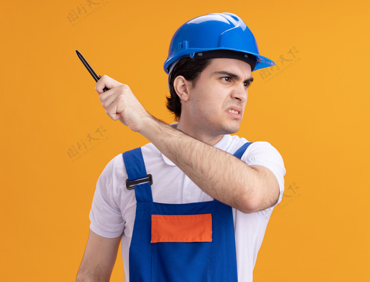 脸年轻的建筑工人穿着建筑制服 戴着安全帽 手里拿着钢笔 愤怒的脸站在橙色的墙上 望向一边拿着安全建筑工人