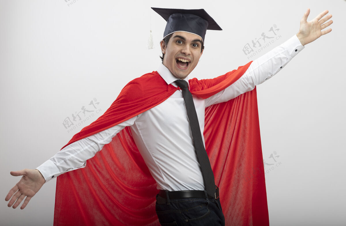 手快乐的年轻超级英雄看着戴着领带和毕业帽的相机 双手分开 背景是白色的传播领带相机
