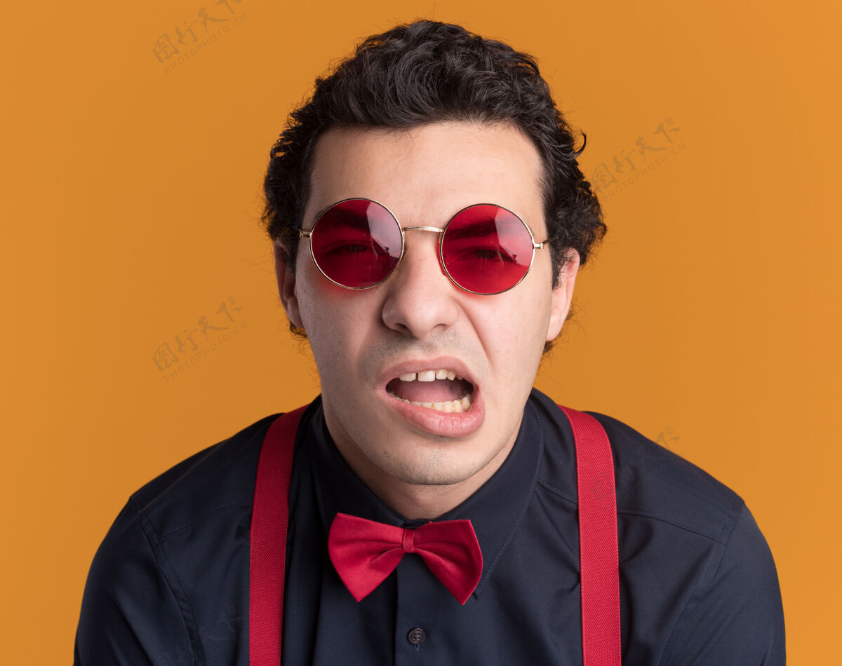 做不满意的时髦男人戴着蝴蝶结戴着眼镜和吊带看着前面 歪着嘴站在橙色的墙上时髦不满意嘴巴