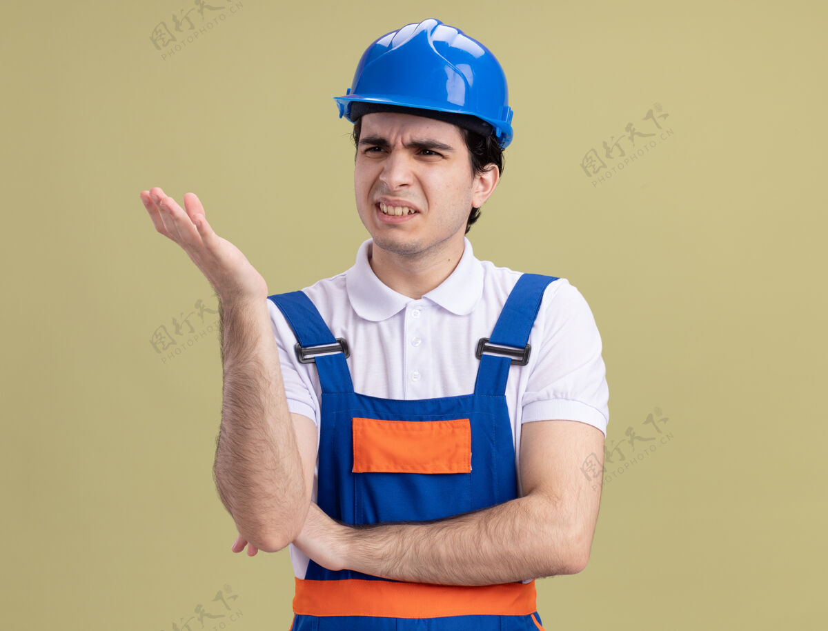 建筑不满意的年轻建筑工人穿着建筑制服 戴着安全帽 站在绿色的墙上 一边看着一边 一边举手表示不满和愤怒制服举起来建筑工人