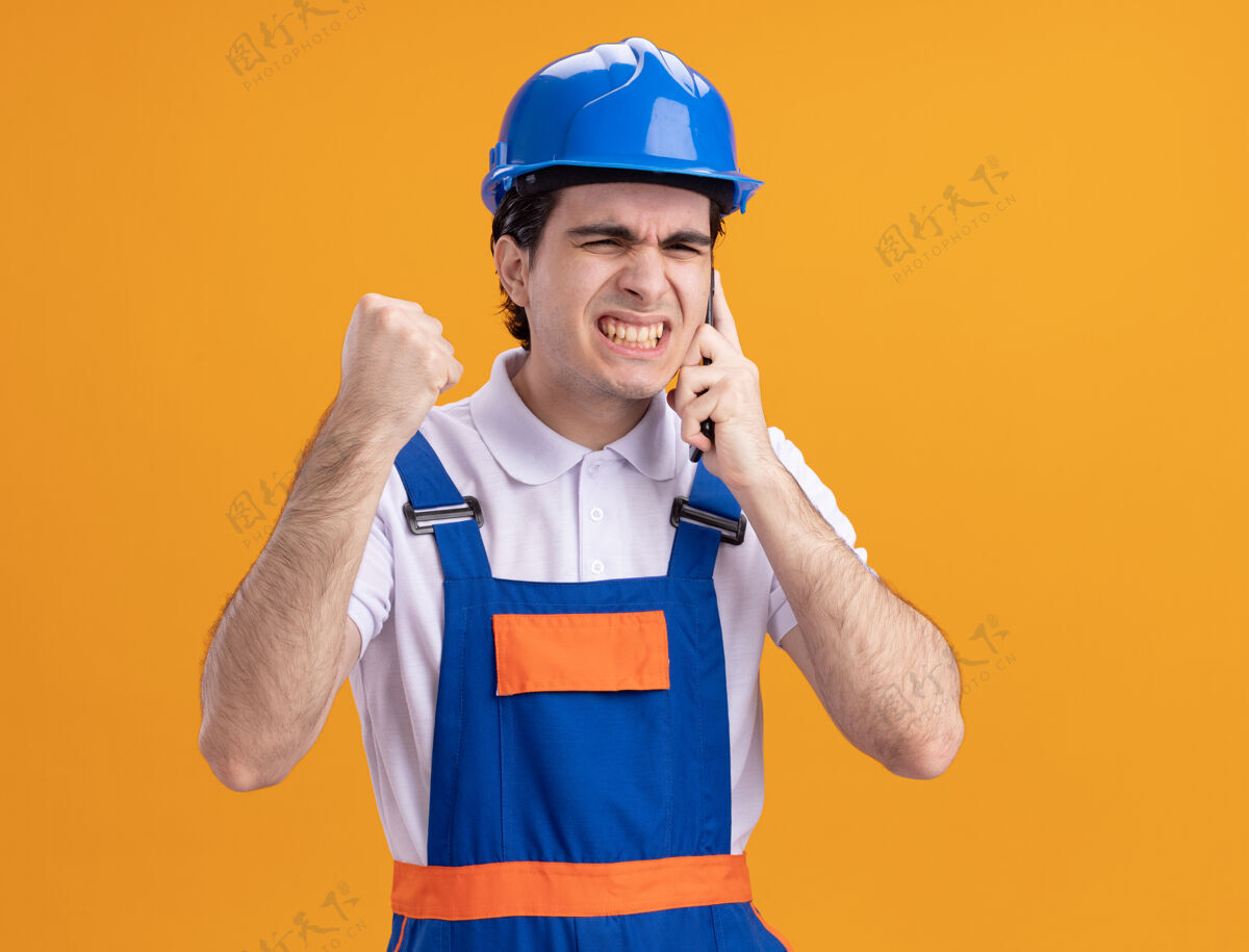 建筑工人愤怒的年轻建筑工人穿着建筑制服 戴着安全帽 站在橙色的墙上讲着手机愤怒男人站着