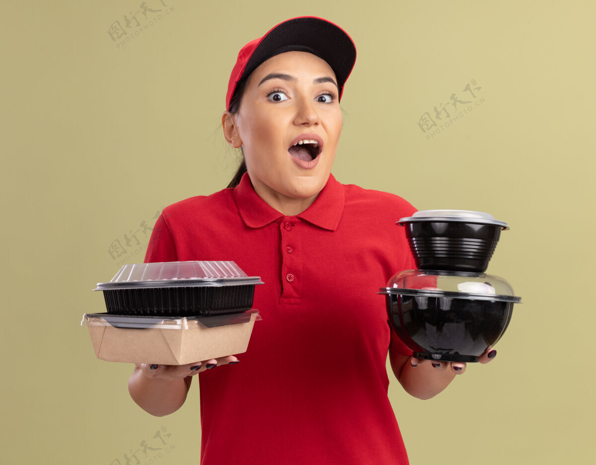 盒子快乐的年轻送货员身穿红色制服 戴着帽子 手里拿着食品包 站在绿色的墙边 快乐而惊讶地看着前面送货食物快乐