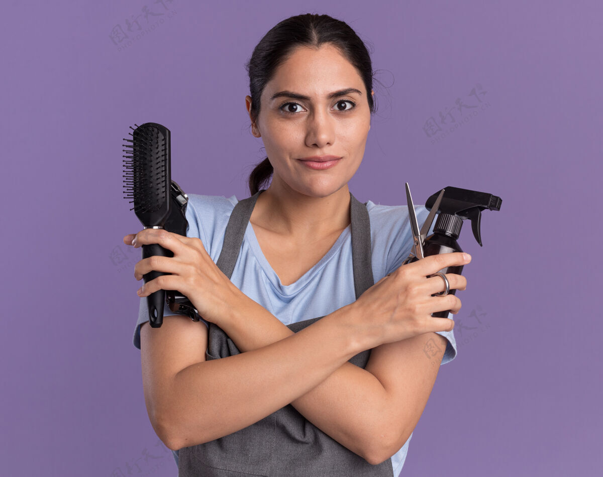 女人穿着围裙的年轻美女理发师拿着喷雾瓶修剪机和毛刷站在紫色的墙上 自信地看着前面修剪者站着美丽