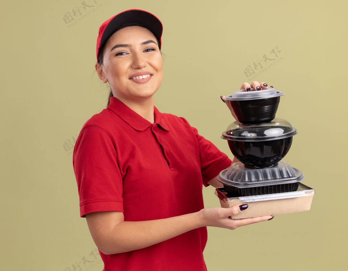 站着身着红色制服 戴着帽子的快乐的年轻女送货员 手里拿着一叠食品包 面带微笑地站在绿色的墙边看着前方年轻叠着快乐