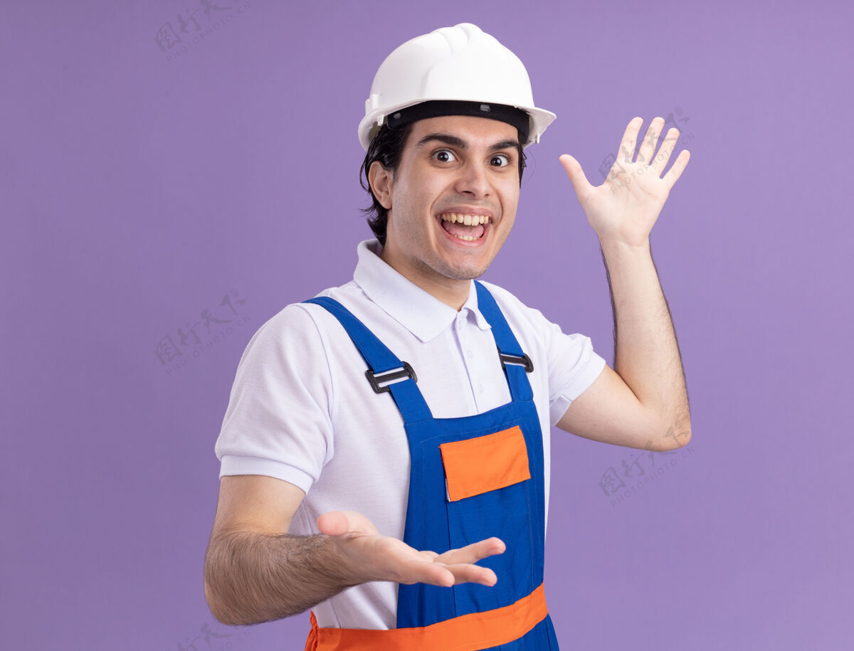 快乐身着施工制服 头戴安全帽的快乐的年轻建筑工人站在紫色的墙壁上 双手打着手势 愉快地微笑着建筑手制服