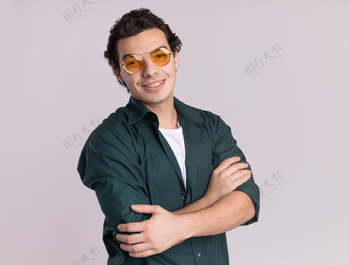 男人穿绿衬衫的年轻人戴着眼镜 面带微笑 双臂交叉着站在白墙上年轻人脸衬衫