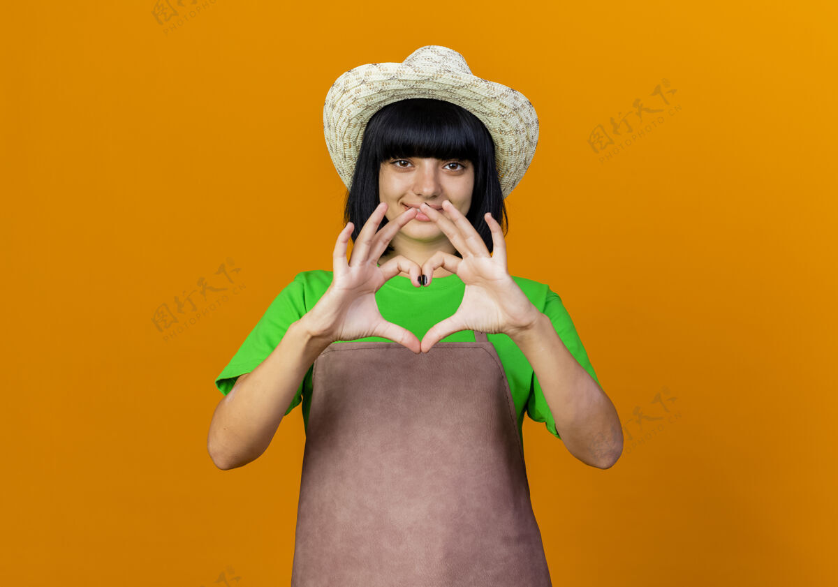 橙色微笑着的年轻女园丁身着制服戴着园艺帽手势心手示意标志穿着手势