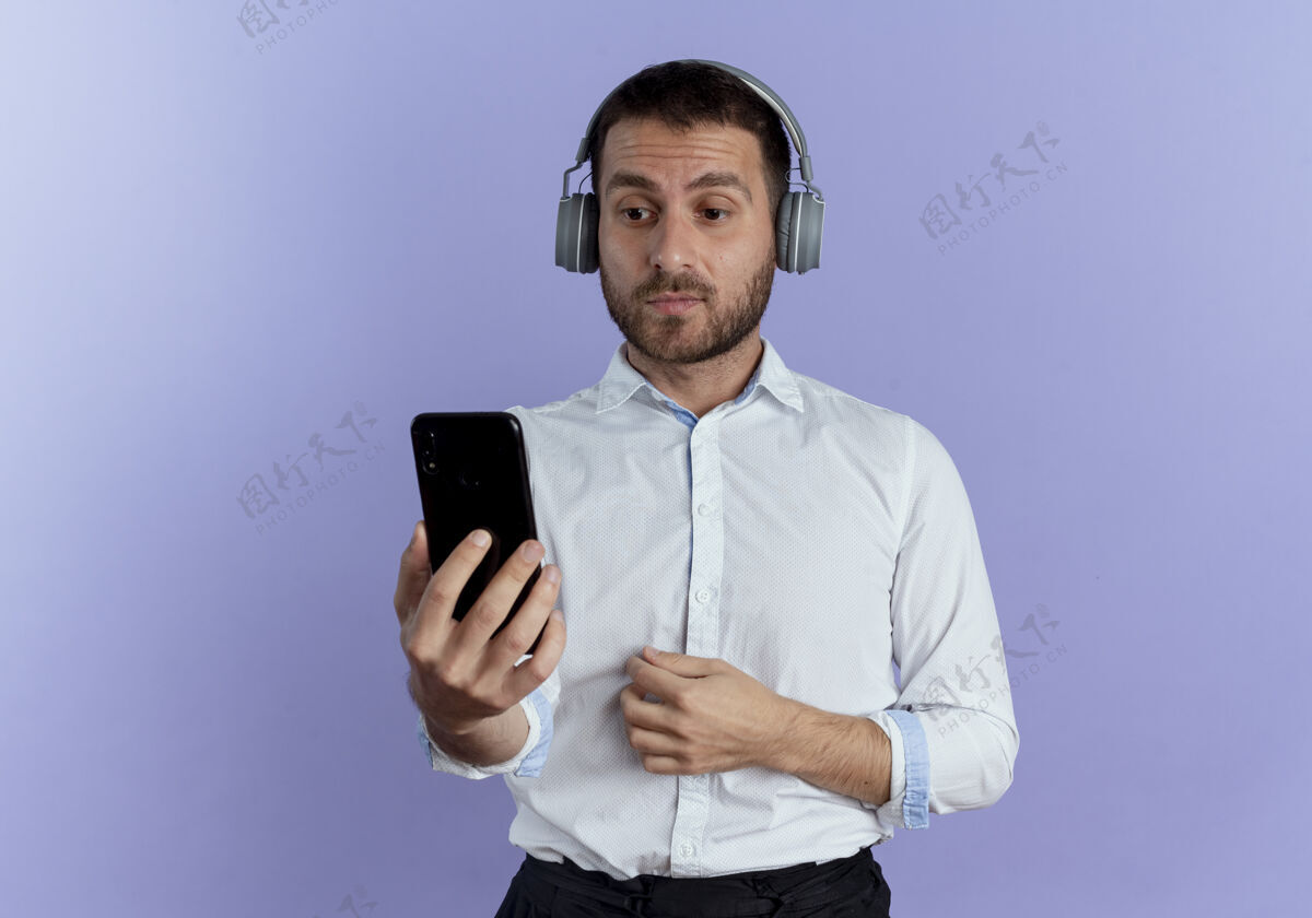 电话戴着耳机的帅哥惊讶地拿着手机 看着隔离在紫色墙上的手机成人优雅年轻
