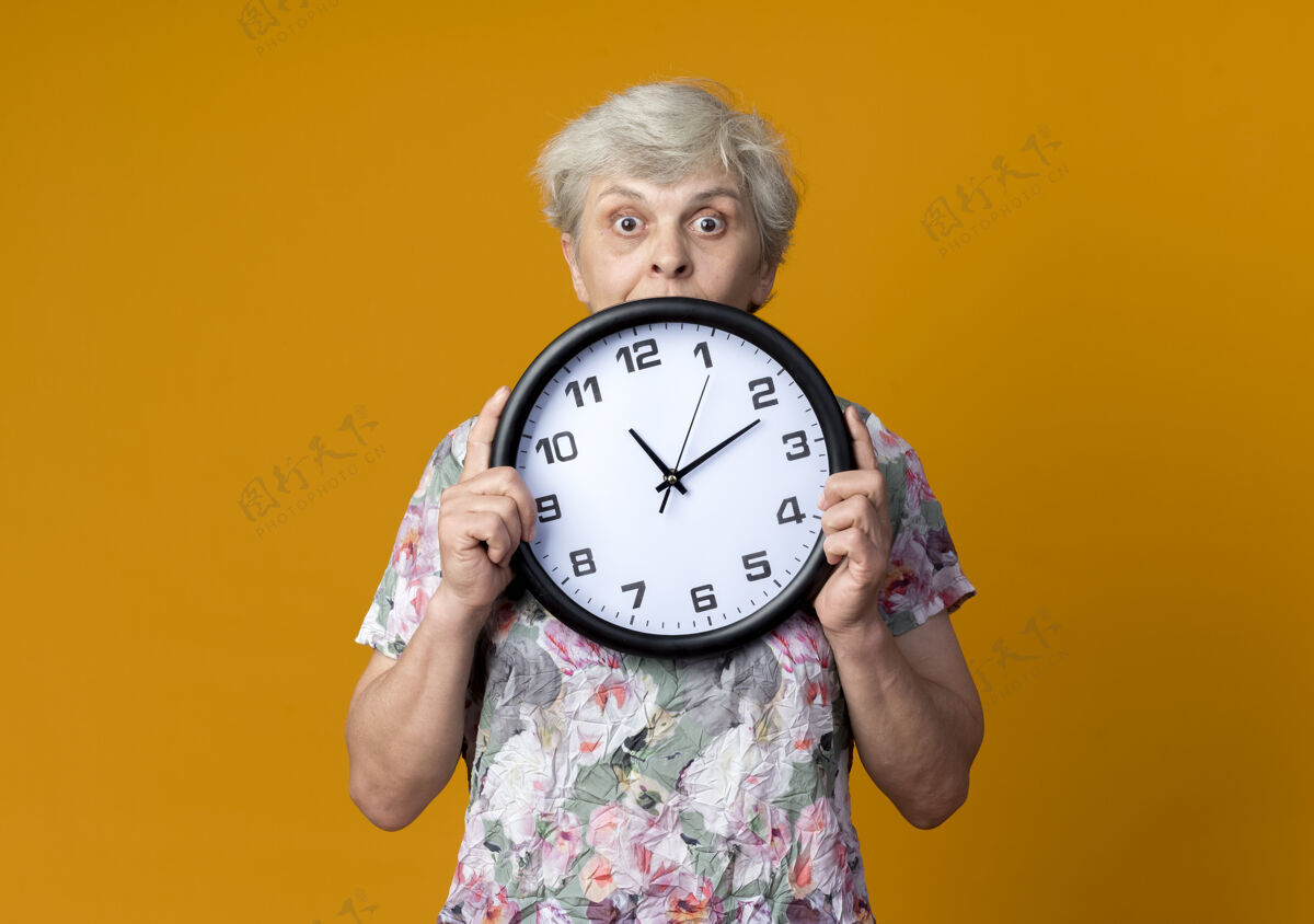 举行惊讶的老妇人拿着并看着隔离在橙色墙上的钟惊喜时钟老人
