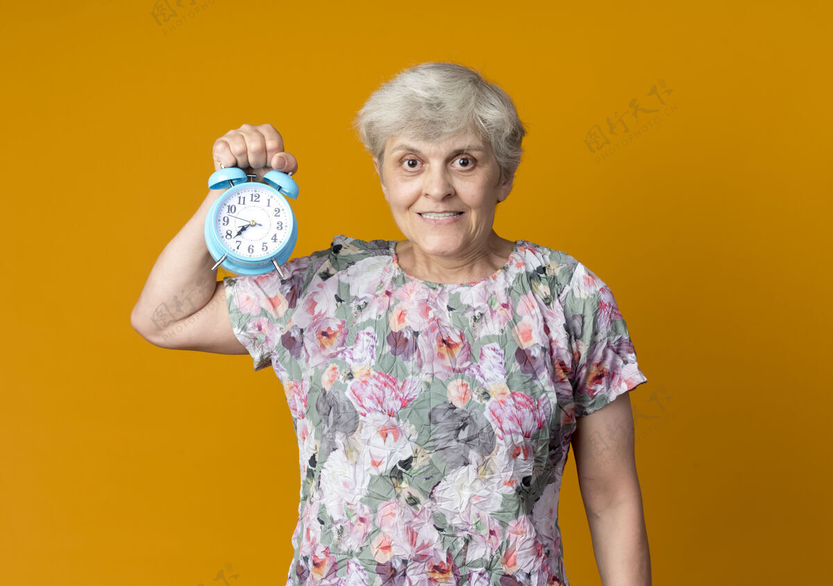抱着微笑的老妇人把闹钟孤立地放在橙色的墙上老人钟老人