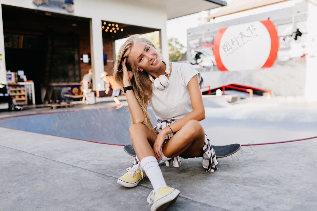活动可爱的白种女人在溜冰场愉快地摆姿势迷人的金发女郎坐在滑板上微笑着配件运动街道