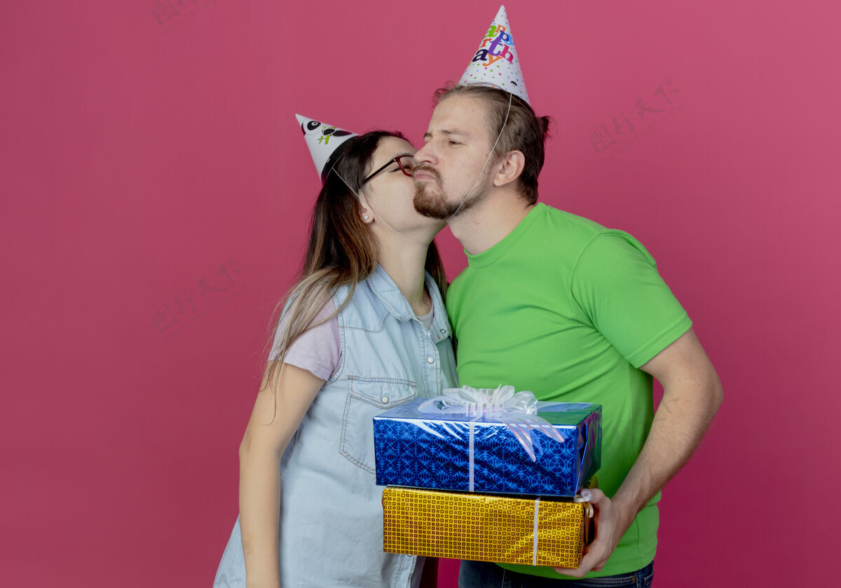 戴着高兴的年轻夫妇戴着聚会帽女孩亲吻男子拿着礼品盒隔离在粉红色的墙上请帽子男人