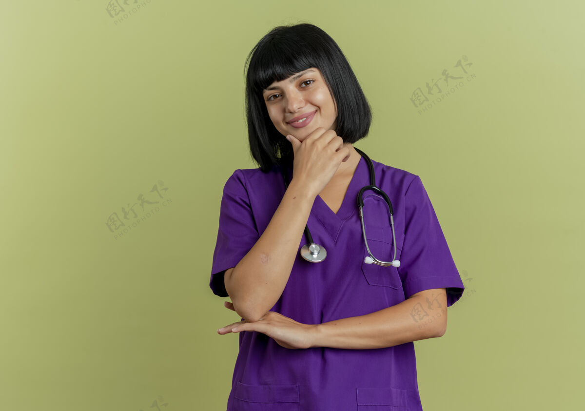背景年轻的黑发女医生穿着制服 手持听诊器 手放在下巴上 看着隔离在橄榄绿背景和复印空间上的相机年轻女性医生