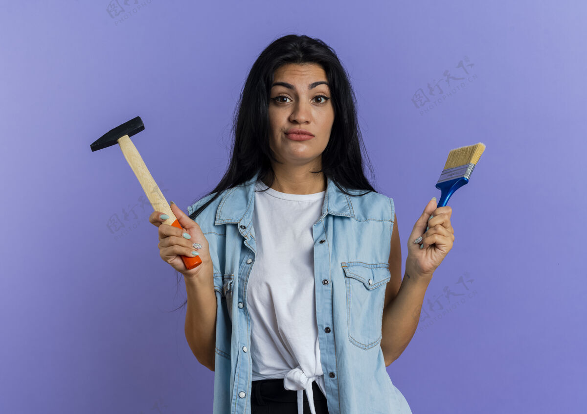 锤子困惑的年轻白种女人拿着锤子和画笔在紫色背景上孤立地复制空间抱着年轻刷子