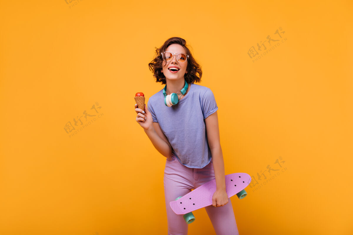 音乐穿着休闲服的漂亮白人女孩戴着耳机摆姿势穿着滑板吃冰淇淋的漂亮女士的画像休闲滑板欢呼