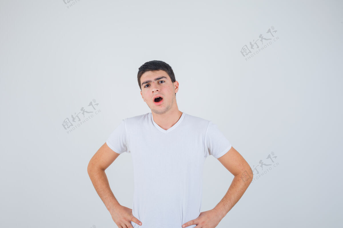 腰年轻的男性手放在腰上 穿着t恤张开嘴 看起来很困惑 正面视图模特态度胡须