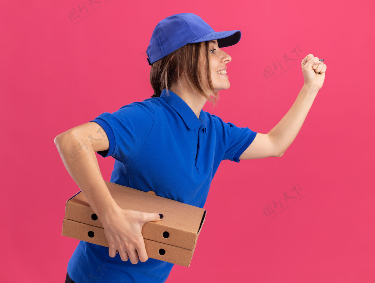 请穿着制服的年轻漂亮的送货女孩站在一边 手里拿着披萨盒 假装是粉红色的跑步披萨制服