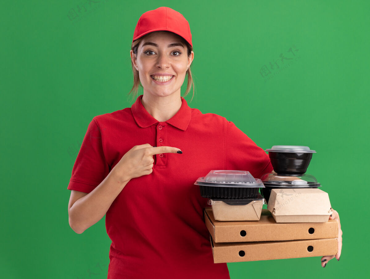 姿势微笑着的年轻漂亮的女送货员穿着制服 指着绿色的比萨饼盒上的纸食品包装和容器漂亮送货成人