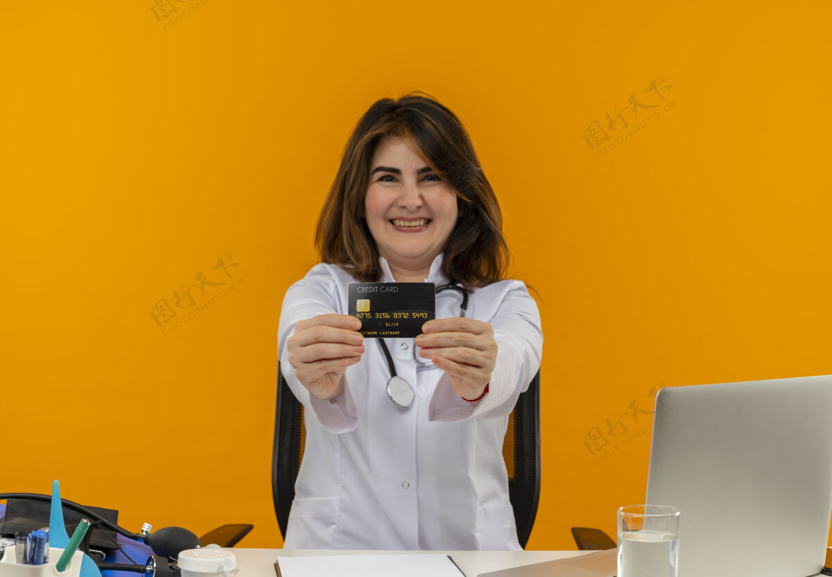 工具快乐的中年女医生穿着医用长袍和听诊器坐在办公桌旁 用医疗工具剪贴板和笔记本电脑伸开信用卡隔离伸展医生快乐