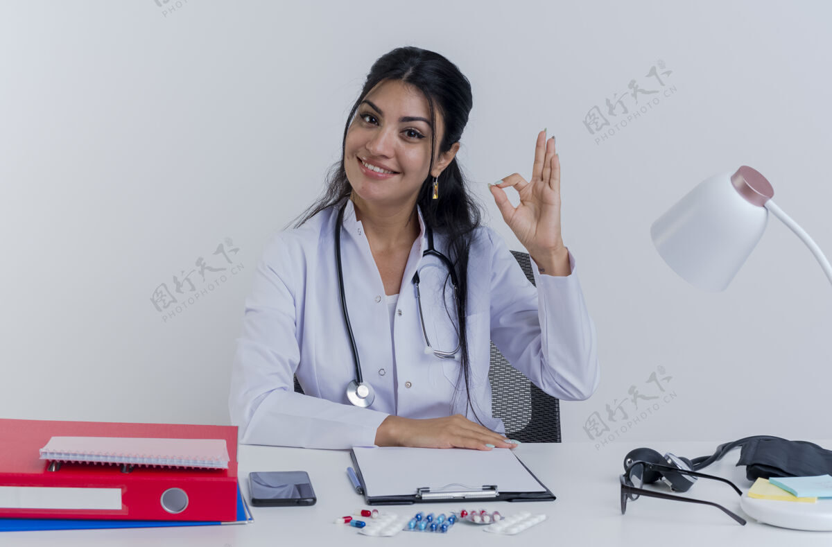 桌子面带微笑的年轻女医生穿着医用长袍和听诊器坐在办公桌旁拿着医疗工具看着把手放在办公桌上做着ok标志隔离成年人医疗医生
