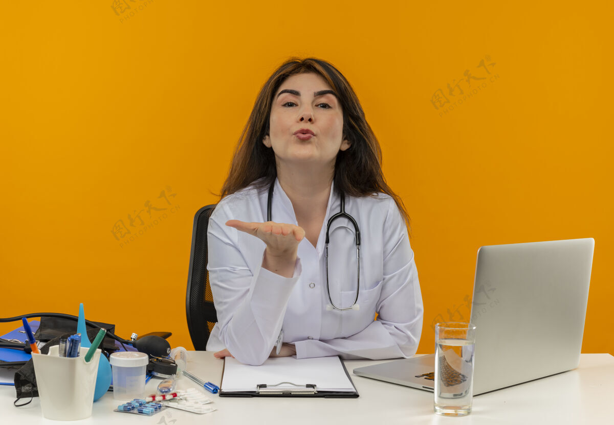 中年自信的中年女医生穿着医用长袍和听诊器坐在办公桌旁 拿着医疗工具 剪贴板和笔记本电脑 看着和送人吹吻发送女性医生