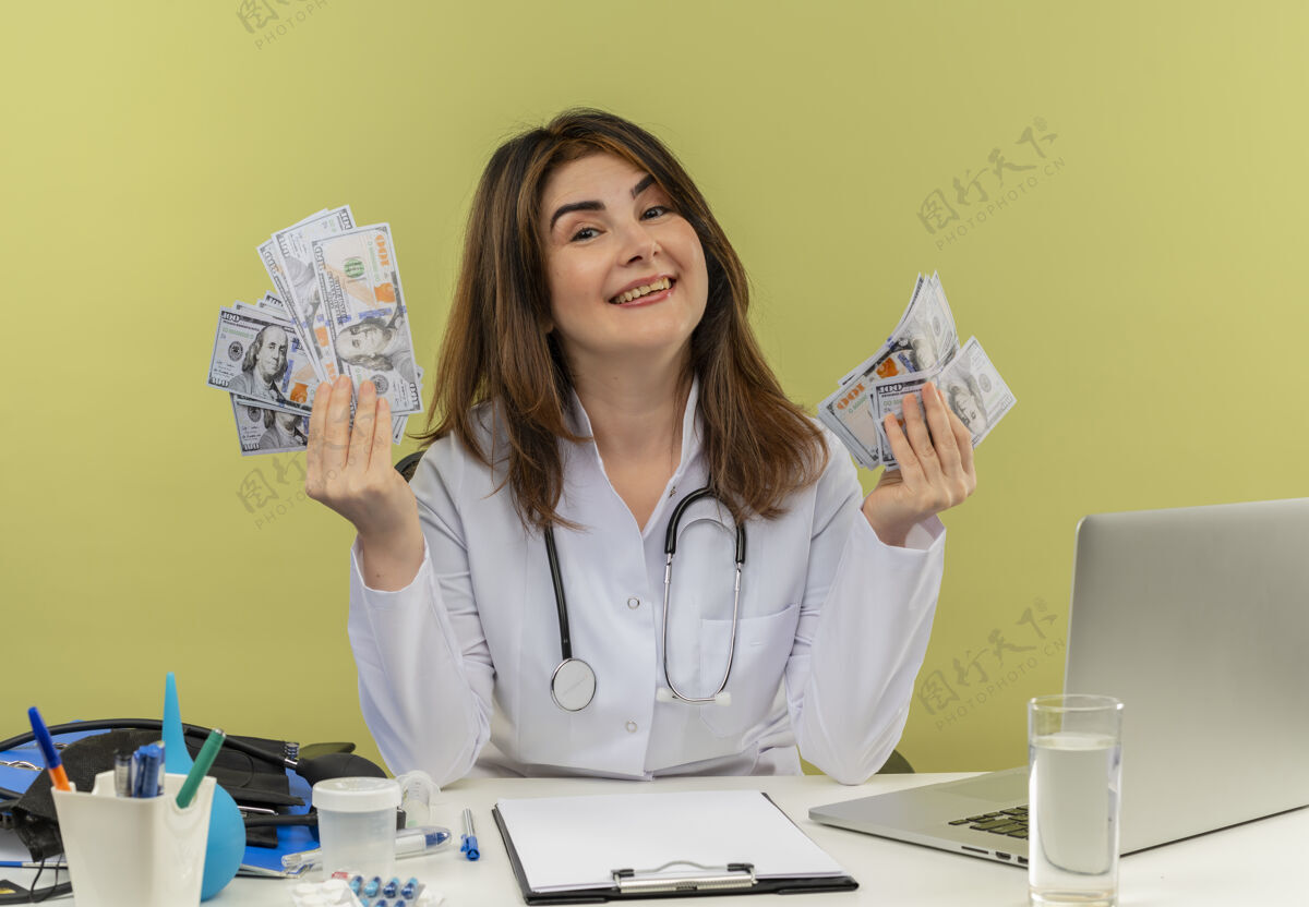 工具微笑的中年女医生穿着医用长袍和听诊器坐在办公桌旁 手里拿着医疗工具和笔记本电脑 手里拿着钱 看上去与世隔绝坐着微笑成人