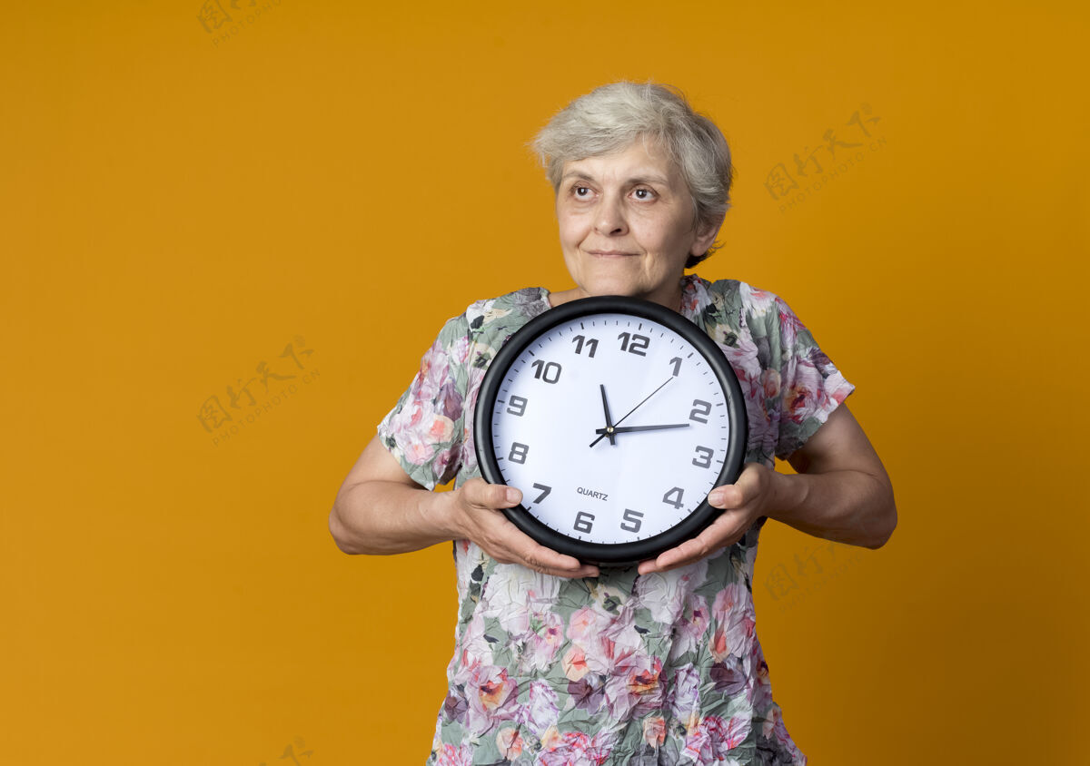 老人高兴的老太婆拿着钟看着橘色墙上孤立的一面请等一下橙色时钟
