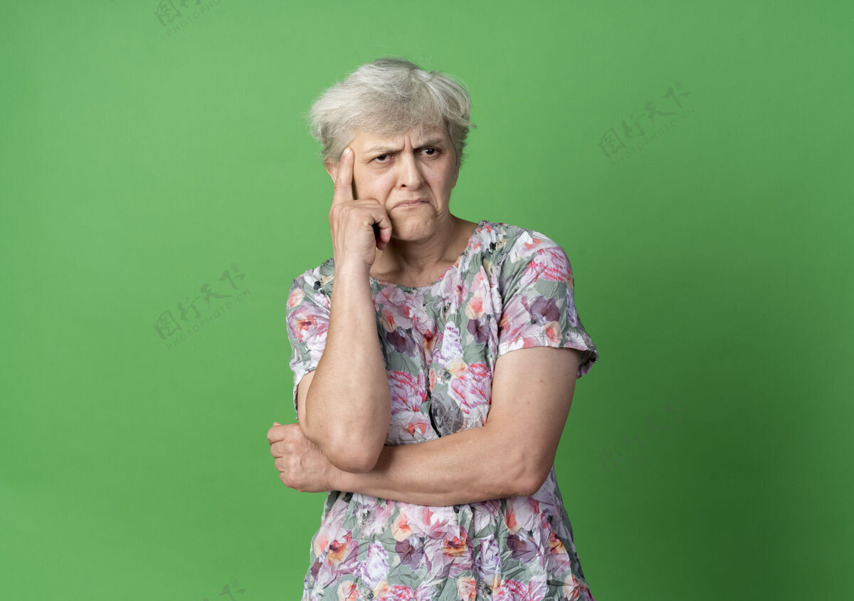 绿的困惑的老妇人把手放在下巴上 看着隔离在绿墙上的一面女人老的手