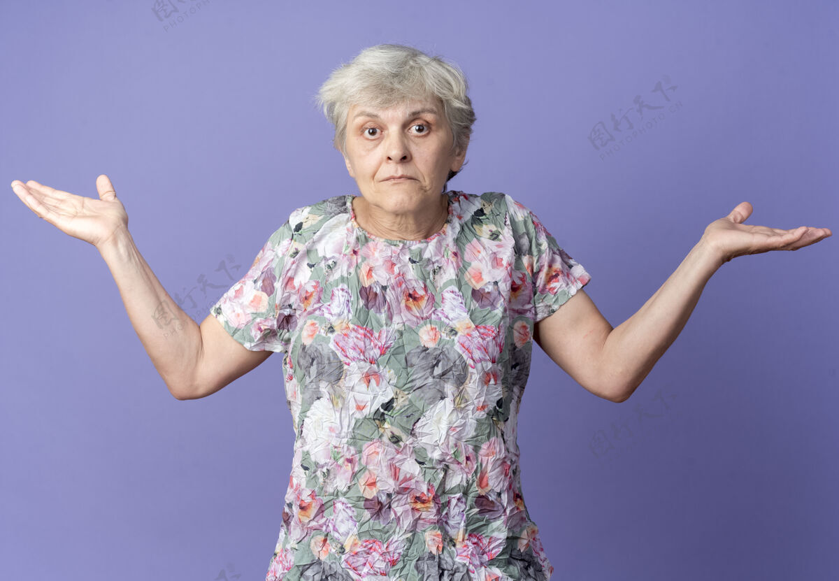 老困惑的老妇人站在紫色的墙上 双手张开女人老手