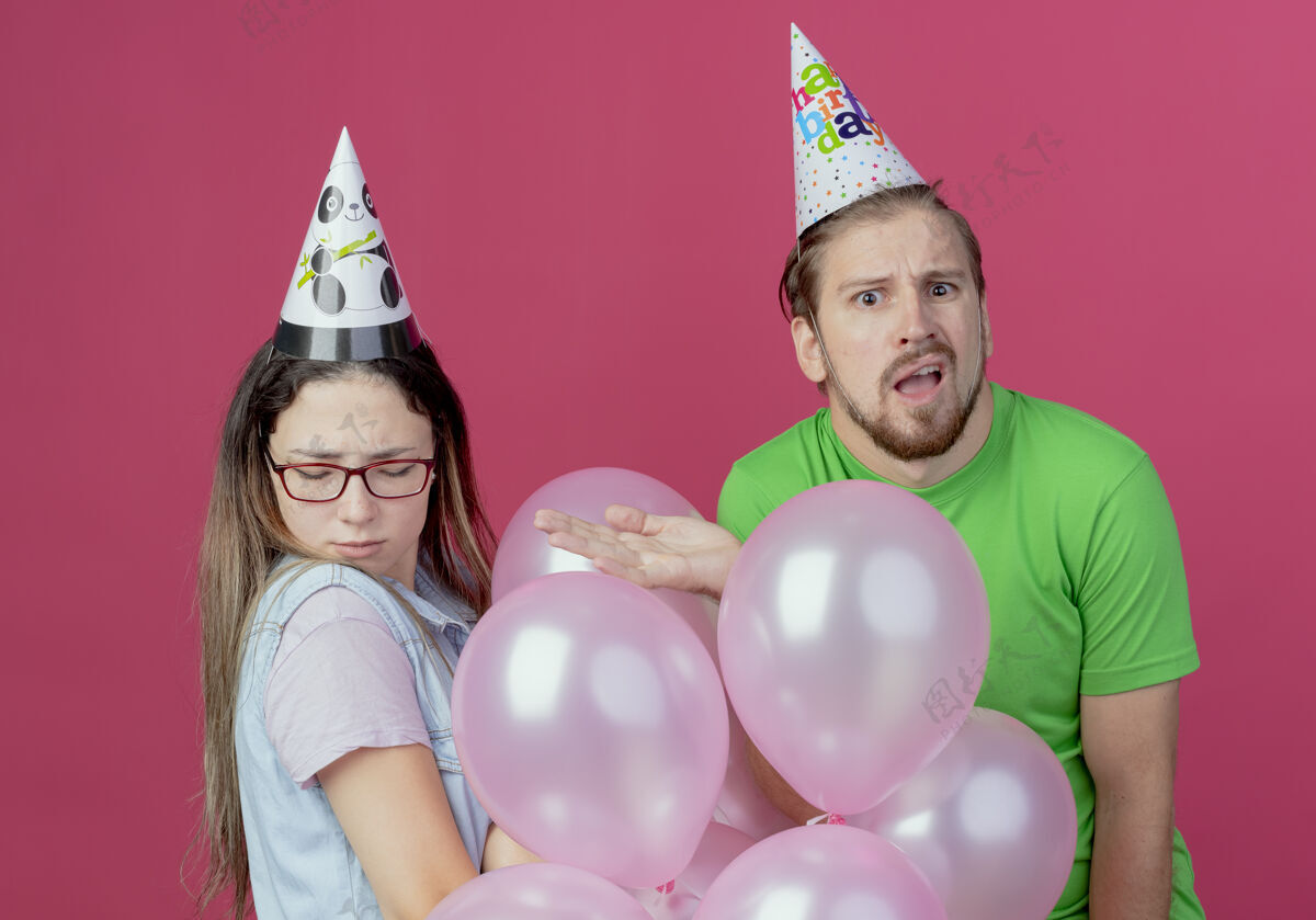 聚会戴着派对礼帽的困惑的年轻人指着站在粉红色墙上的氦气球的生气的年轻女孩粉色生日年轻