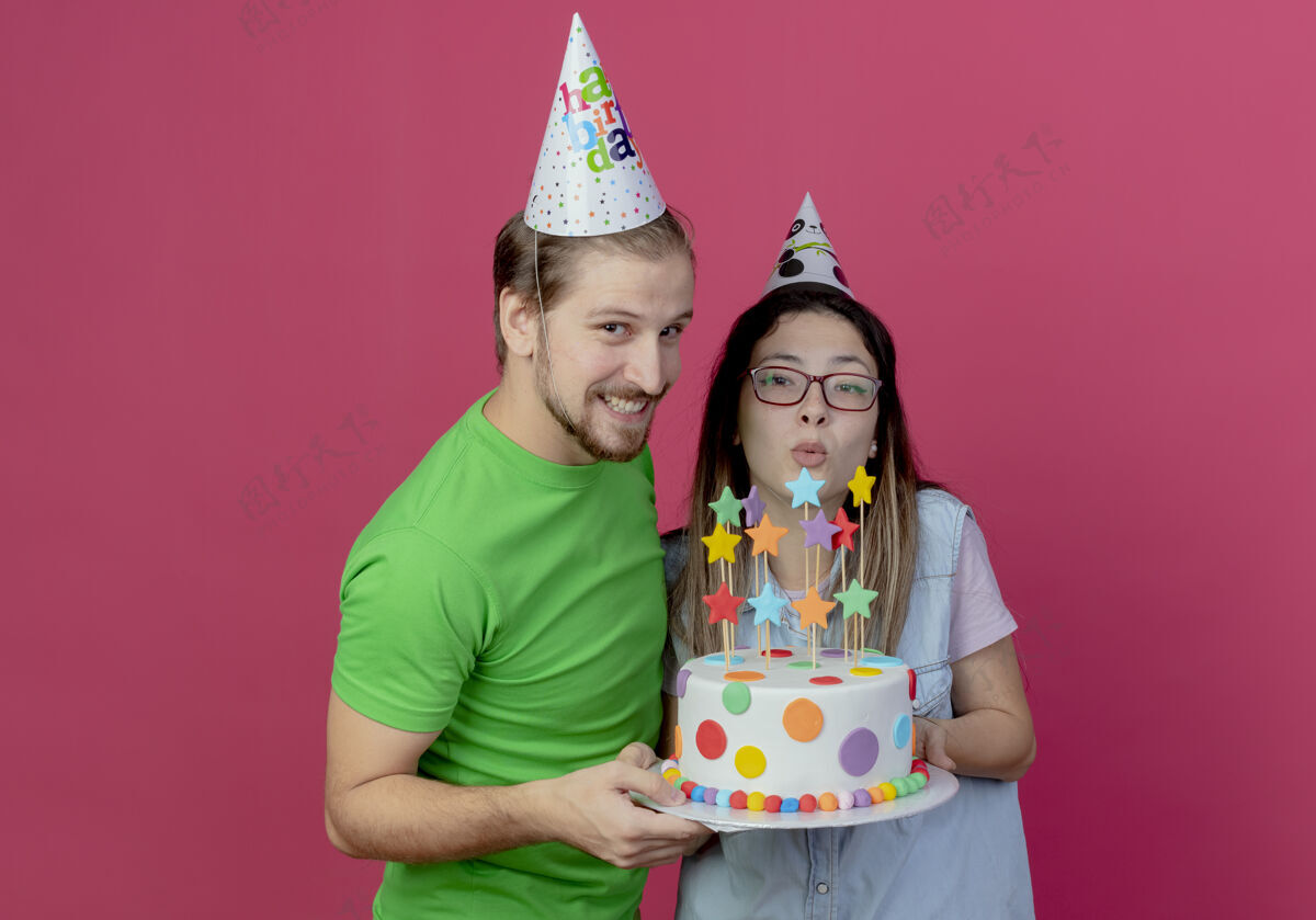 生日快乐的年轻夫妇戴着派对帽 拿着生日蛋糕 看起来孤立地站在粉红色的墙上粉色情侣穿