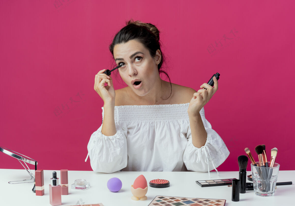 桌子快乐美丽的女孩坐在桌旁 用化妆工具涂睫毛膏隔离在粉红色的墙上粉色睫毛膏涂抹