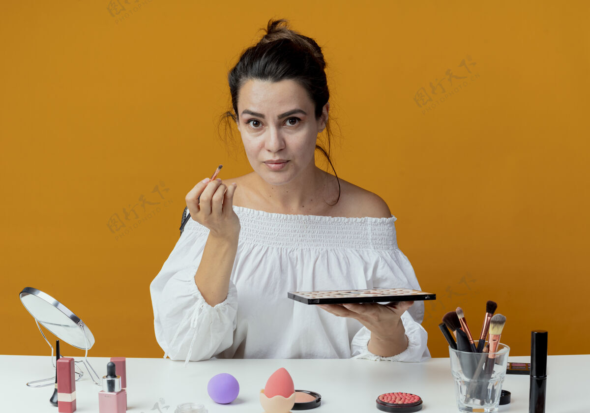 工具美丽的女孩坐在桌子上 用化妆工具拿着化妆刷和眼影调色板 在橙色的墙上看调色板橘子化妆品