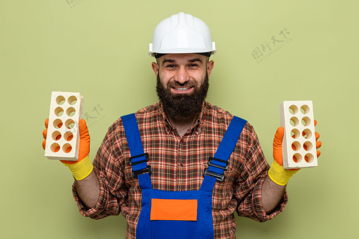 欢呼留着胡须的建筑工人穿着建筑制服 戴着安全帽 戴着橡胶手套 拿着砖头 看着摄像机 微笑着 站在绿色的背景上 快乐而积极胡须手持制服