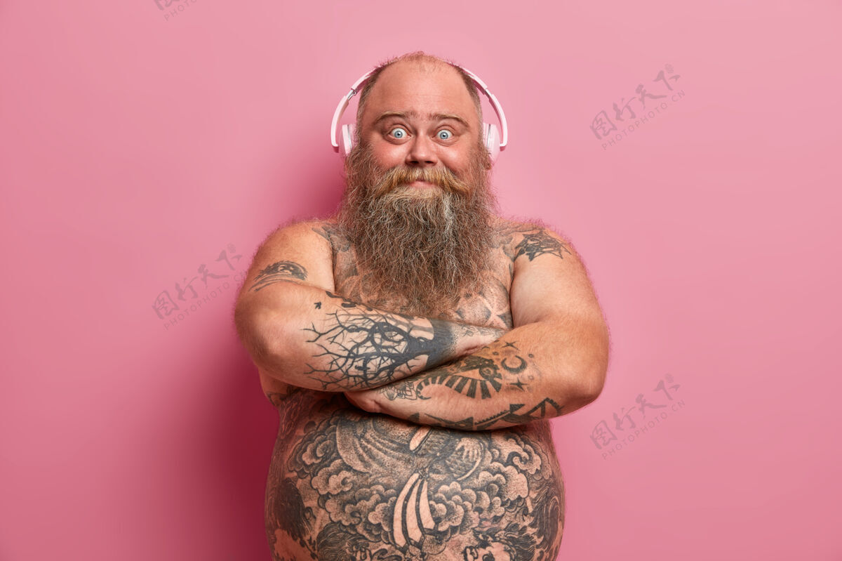 脂肪快乐的胡须男人双手交叉在裸体上 看起来很快乐 喜欢听音乐 耳朵上戴着耳机 肚子上有纹身 听最喜欢的歌家庭休闲 生活方式大丰满胡须