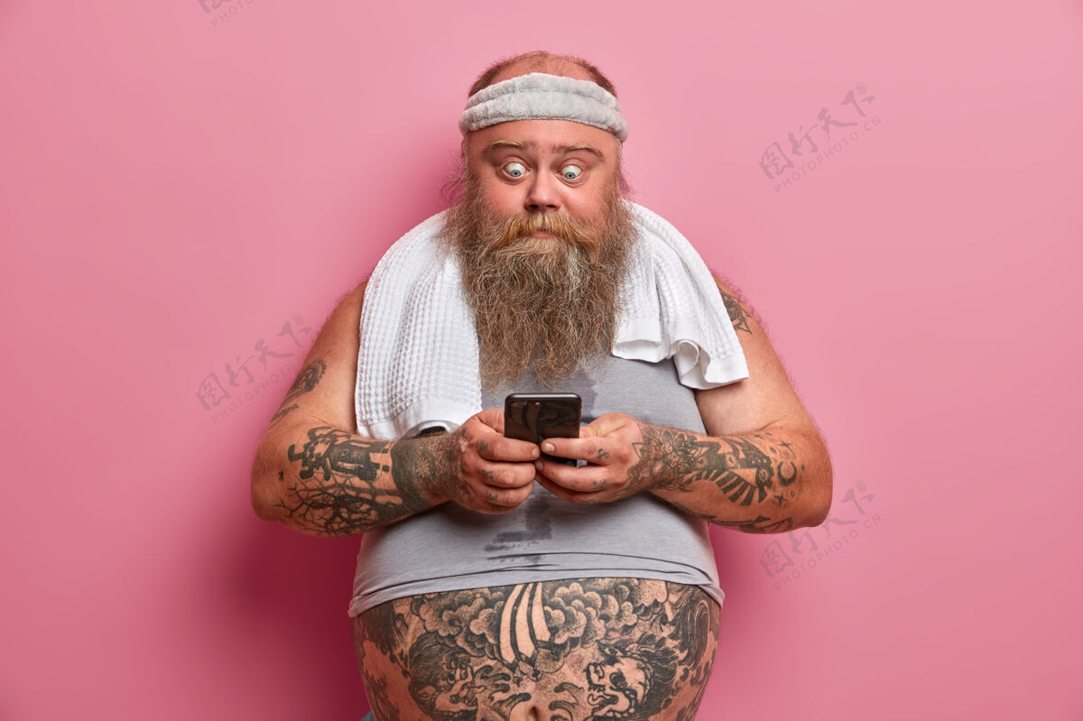 奇迹穿着运动服的大胡子男人 在家锻炼后休息 拿着智能手机 燃烧卡路里和脂肪 下载应用程序 对着玫瑰色的墙壁摆姿势胖胖的家伙带着小玩意儿大怪胎胡须
