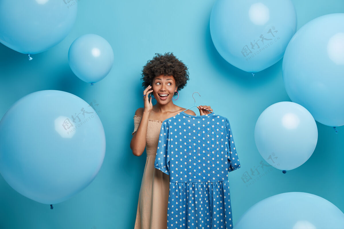 精品快乐的美女把蓝色圆点连衣裙挂在衣架上 给别人打电话 用手机 准备特殊活动 选择服装 在气球周围摆姿势服装 衣柜 时尚概念购物狂购买服装